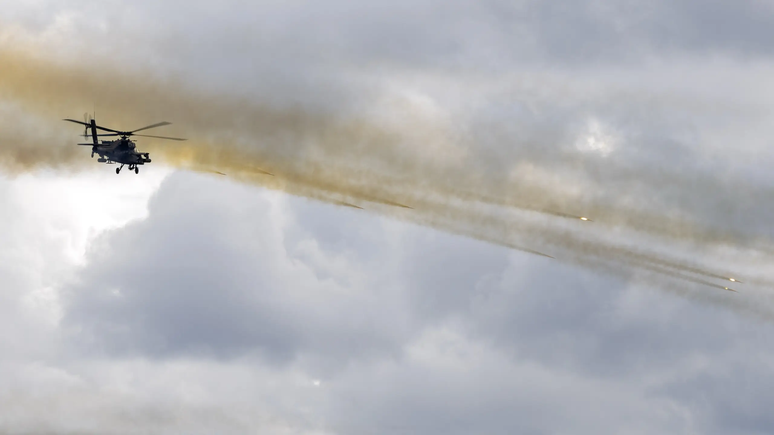 AH-64 Apache firing a salvo of rockets, leaving long smoke trails across a cloudy sky
